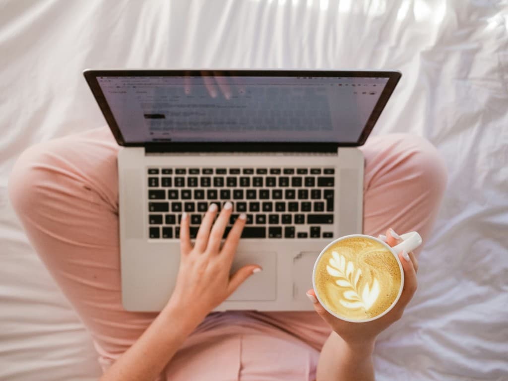 Person working on a laptop with coffee on a white bed